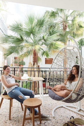 Two people sitting in a hanging chair and on a stool in a courtyard with palm trees and a white table.