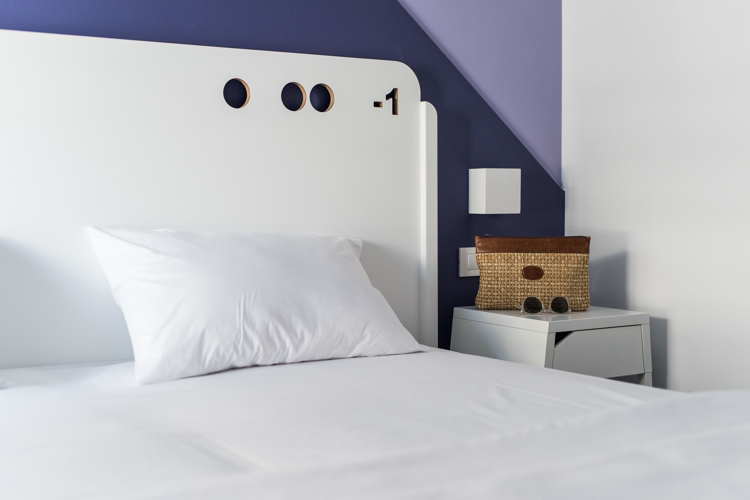 A neatly made bed with white bedding and a single white pillow. A small white nightstand with a woven basket and a lamp is beside the bed.