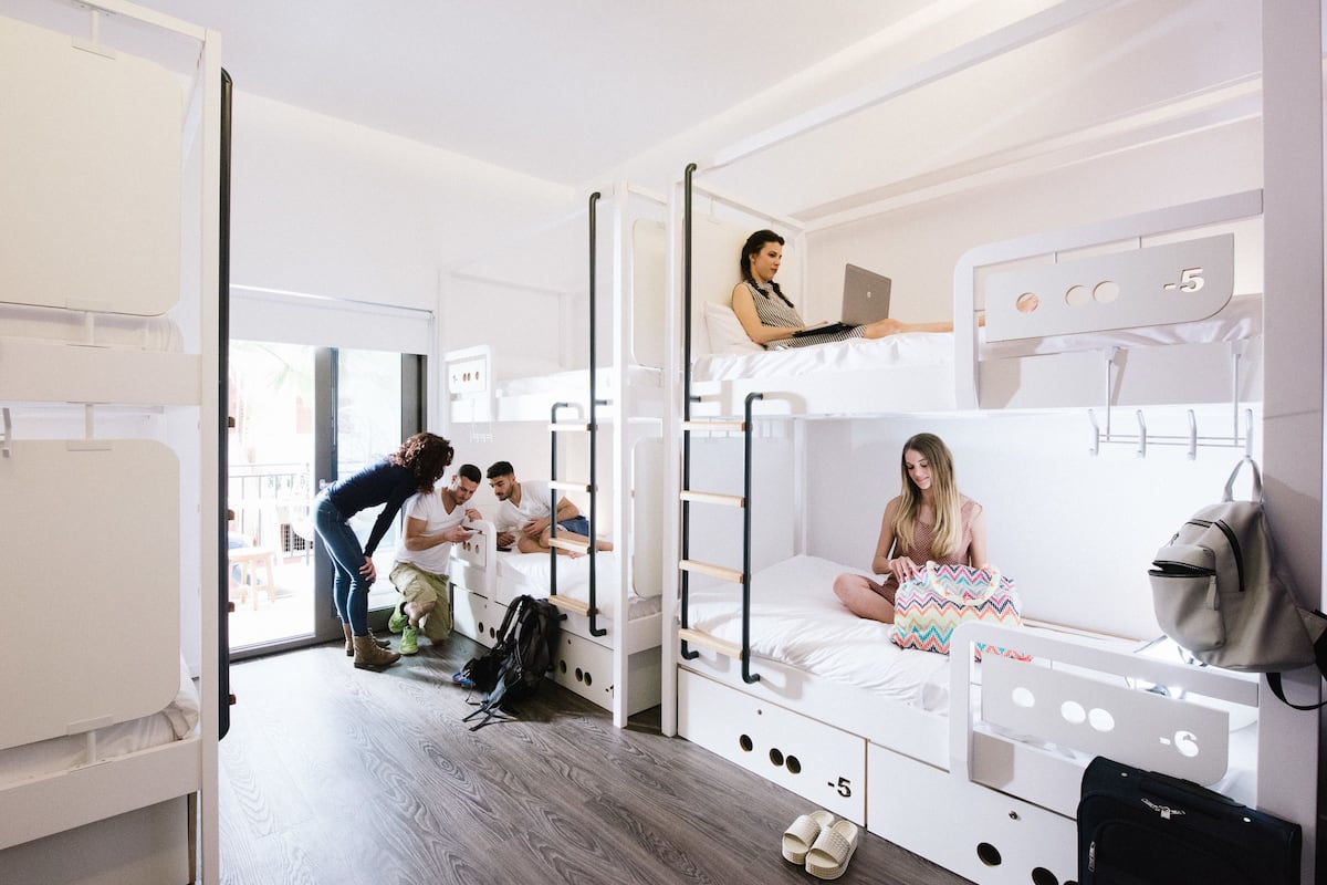Ein Schlafsaalzimmer mit Etagenbetten, ein Laptop auf dem oberen Bett und eine Person, die auf dem unteren Bett sitzt.