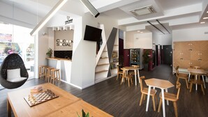 A modern café with a chessboard on a wooden table, a sofa, and a staircase leading to a lower level. - Cocoon City Hostel (Chania)
