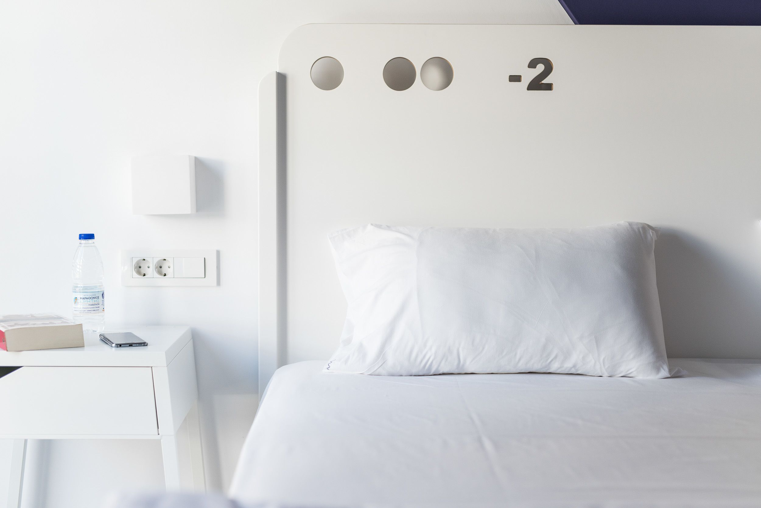 A modern hotel room with a white bed, a nightstand with a water bottle and a book, and a wall-mounted power outlet.