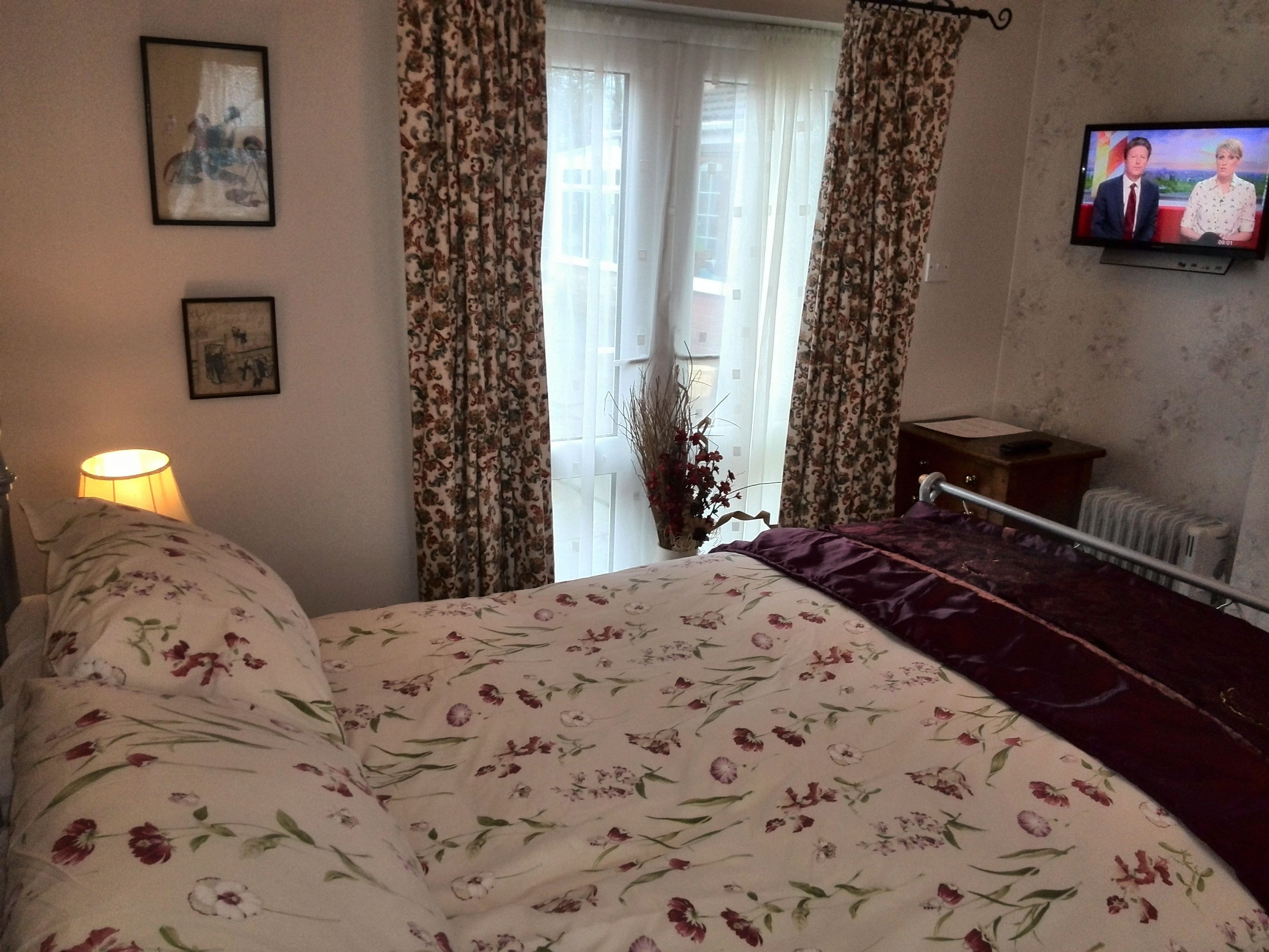 A bedroom with a floral bedspread, a bedside lamp, a television on the wall, and a window with curtains.
