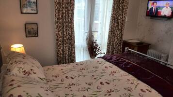 A bedroom with a floral bedspread, a bedside lamp, a television on the wall, and a window with curtains.