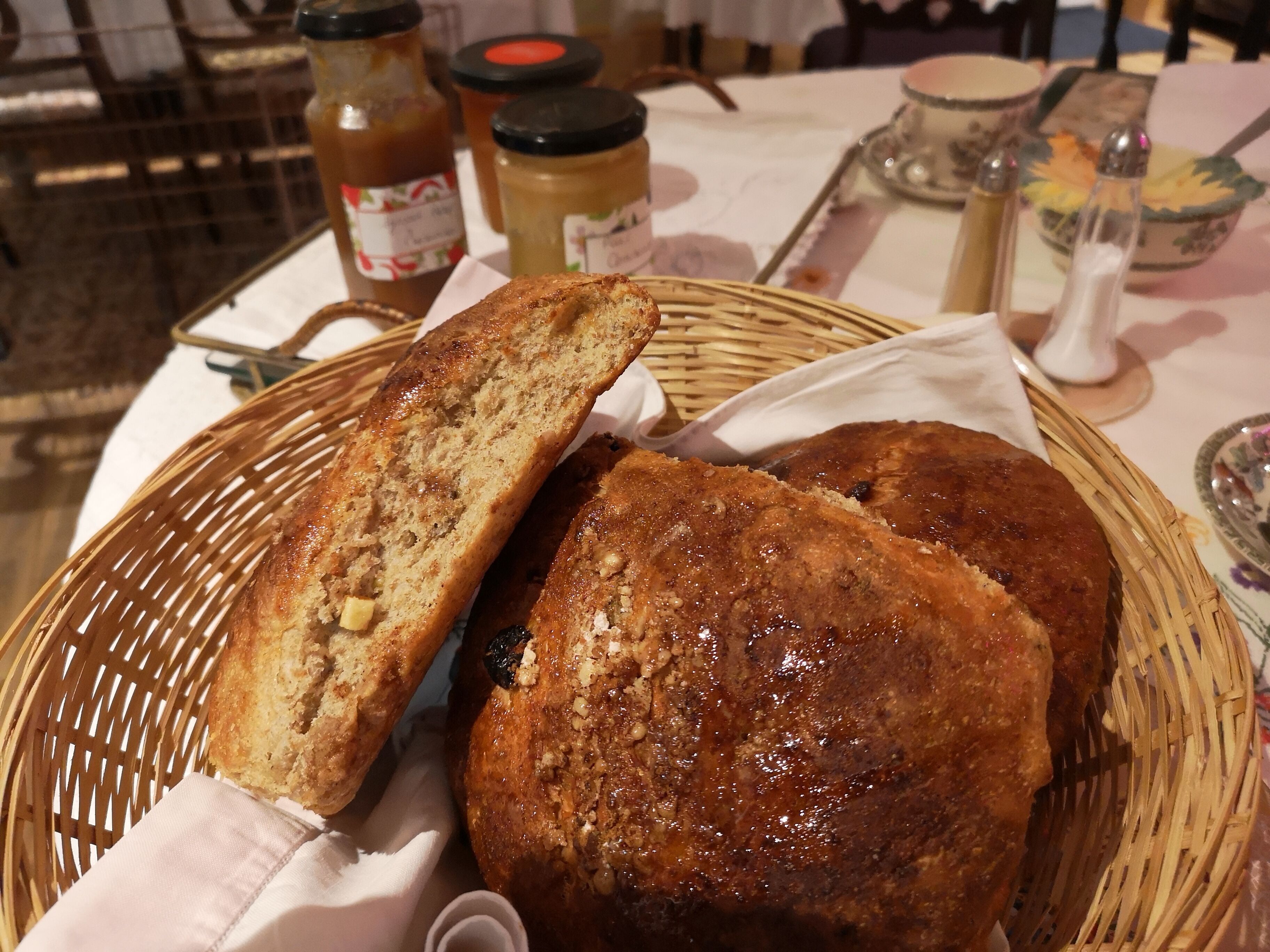 A wicker basket containing two loaves of bread, a jar of jam, and a jar of honey on a table.