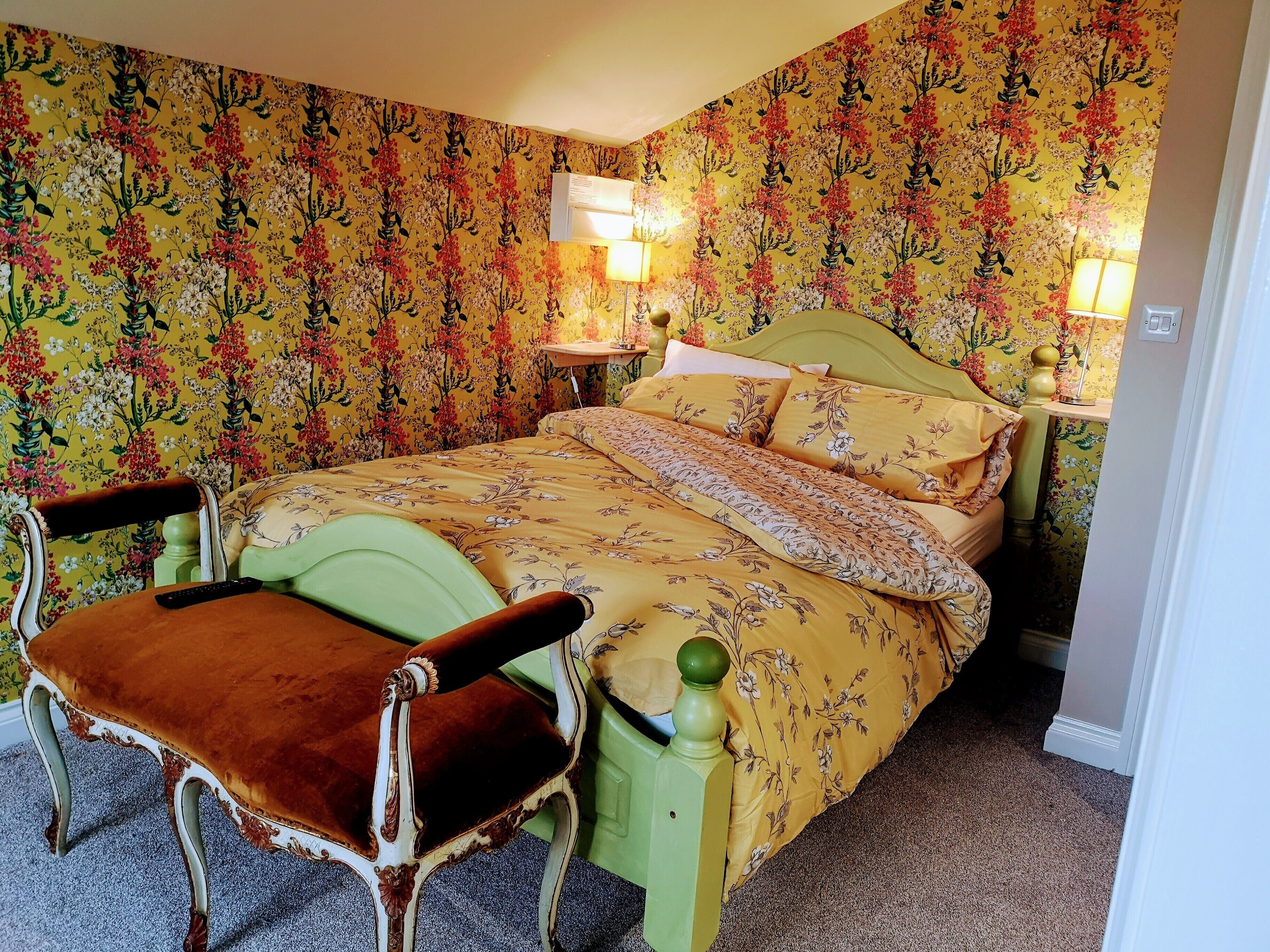 Une chambre avec un papier peint à motifs floraux, un fauteuil recouvert d’un tissu vert, un lit avec une parure assortie à motifs floraux, deux lampes de chevet et une table de chevet.