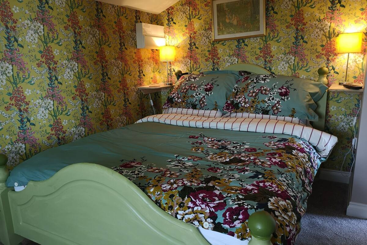 Quarto com papel de parede floral, cama verde, almofadas listradas e duas luminárias de cabeceira.