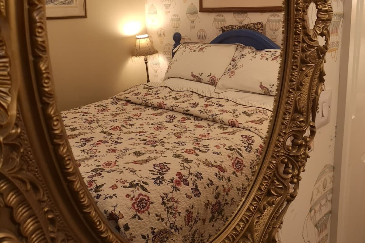 Quarto com um espelho grande e ornamentado, roupa de cama florida, mesa de cabeceira com abajur e papel de parede com desenhos de balões de ar quente.