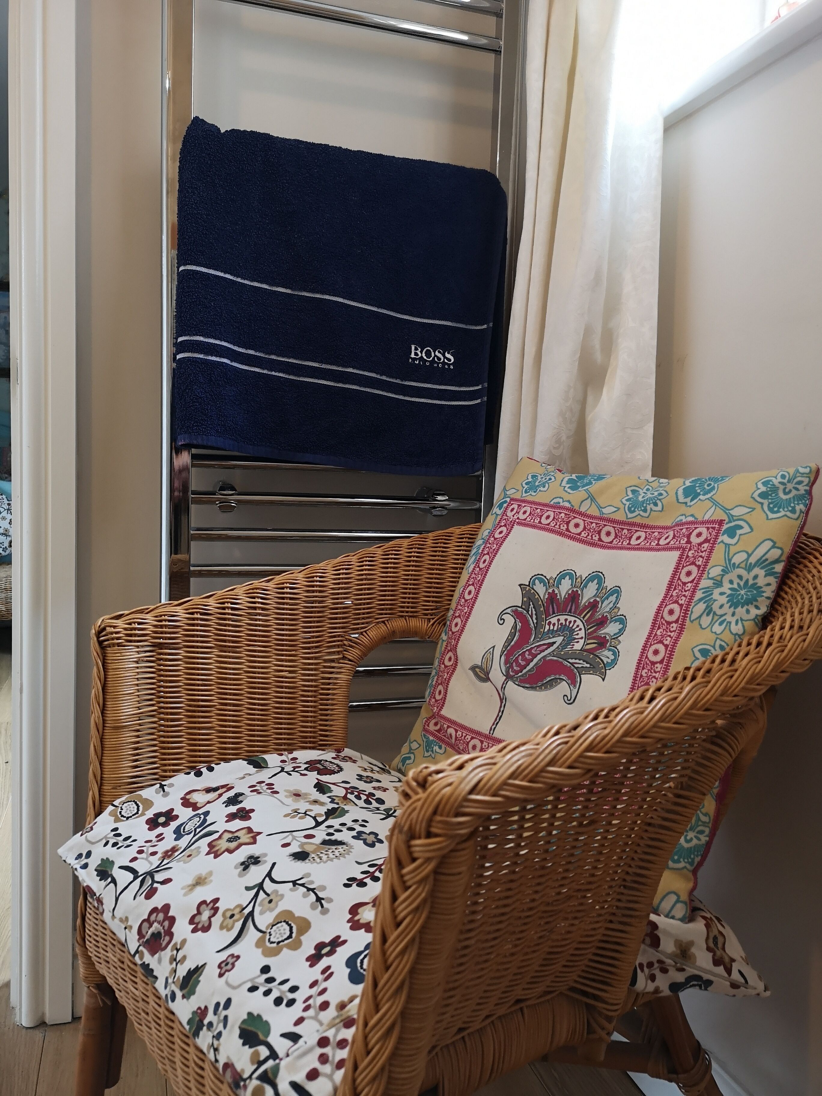 A wicker chair with a floral cushion and a decorative pillow, a metal drying rack with a dark blue towel, and a window with white curtains.
