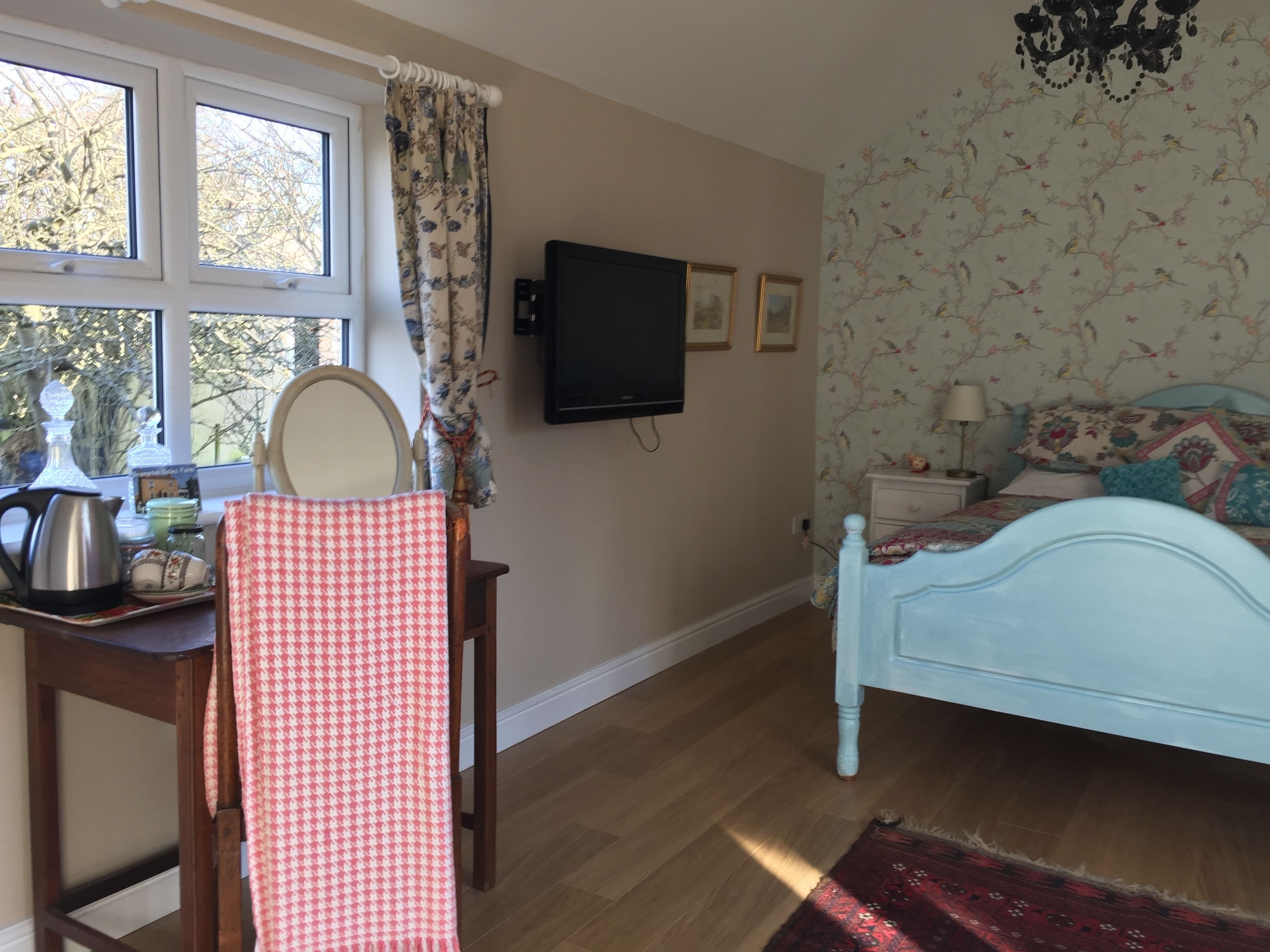 A bedroom with a bed, a TV, a window with curtains, a dresser, and a small table with a kettle and teapot.