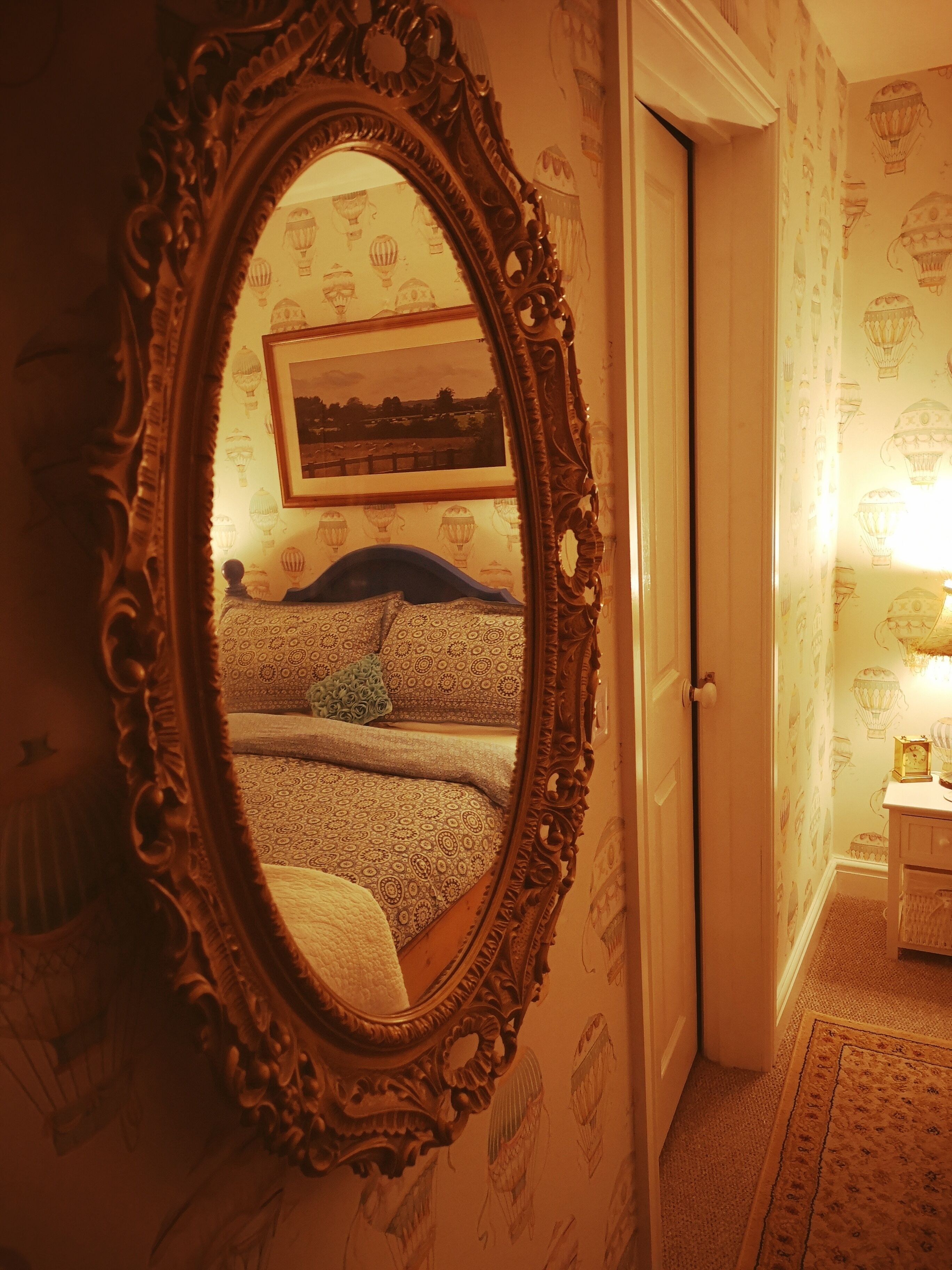 A room with a large ornate mirror reflecting a bed with patterned bedding and a framed landscape painting.