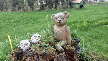 Cena rústica ao ar livre com uma estátua de urso de pedra sentada sobre um toco de árvore, cercada por grama e pequenas plantas.