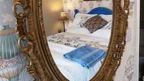 An ornate mirror reflecting a bedroom with patterned wallpaper, a bed with a blue blanket, and a nightstand with a lamp. - Brampton Dales Farm B&B (Gainsborough)