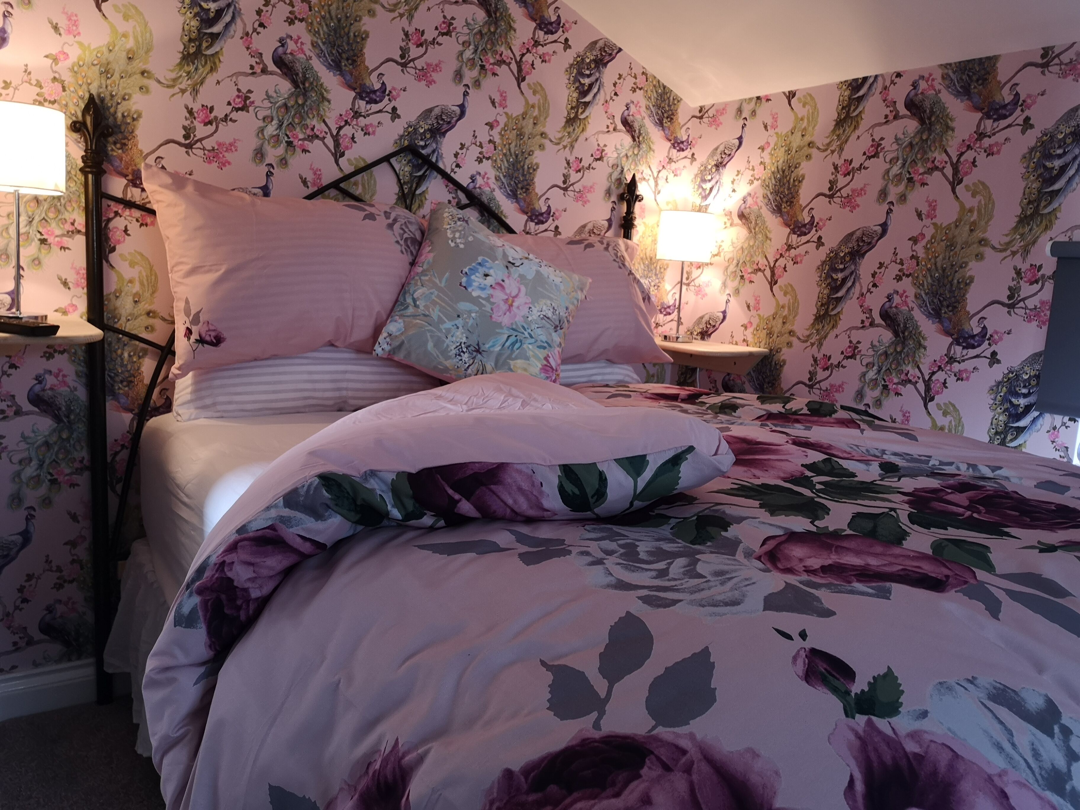 A bedroom with floral wallpaper, a bed with pink bedding, two bedside lamps, and a nightstand with a lamp.
