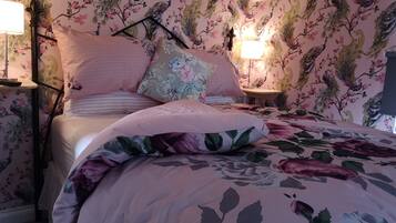 A bedroom with floral wallpaper, a bed with pink bedding, two bedside lamps, and a nightstand with a lamp.