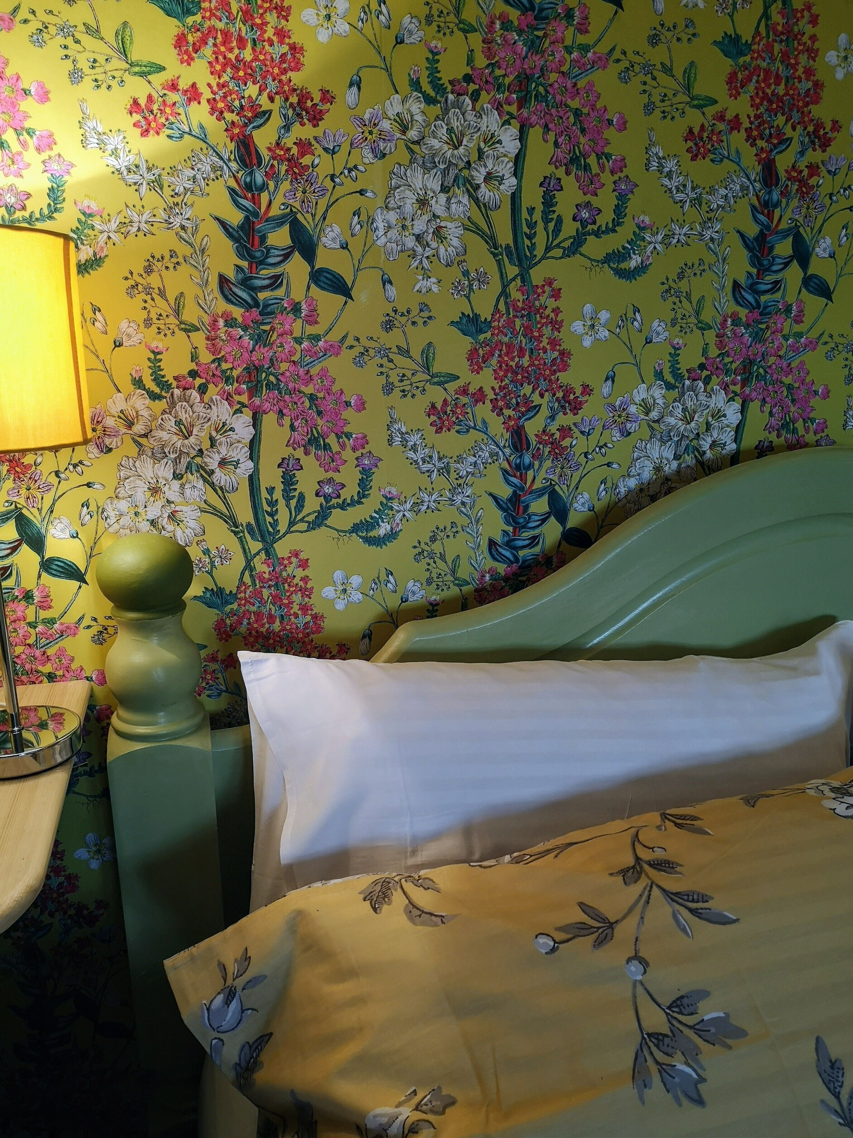 A room with a floral wallpaper, a green bed with white bedding, and a bedside lamp.
