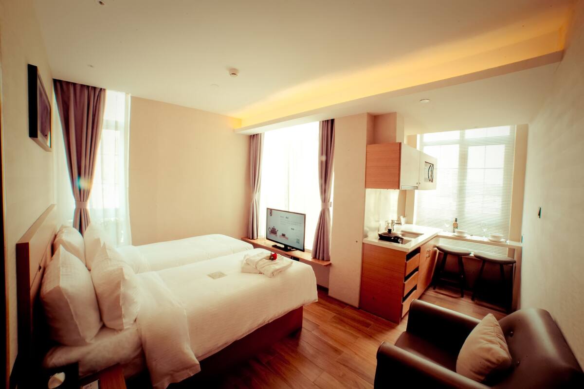 A hotel room with a bed, a desk with a computer, a chair, a small table, and a window with curtains.