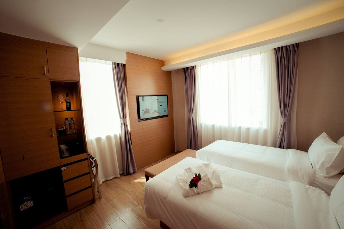 A hotel room with two beds, a wooden wardrobe, a television, and a window with curtains.