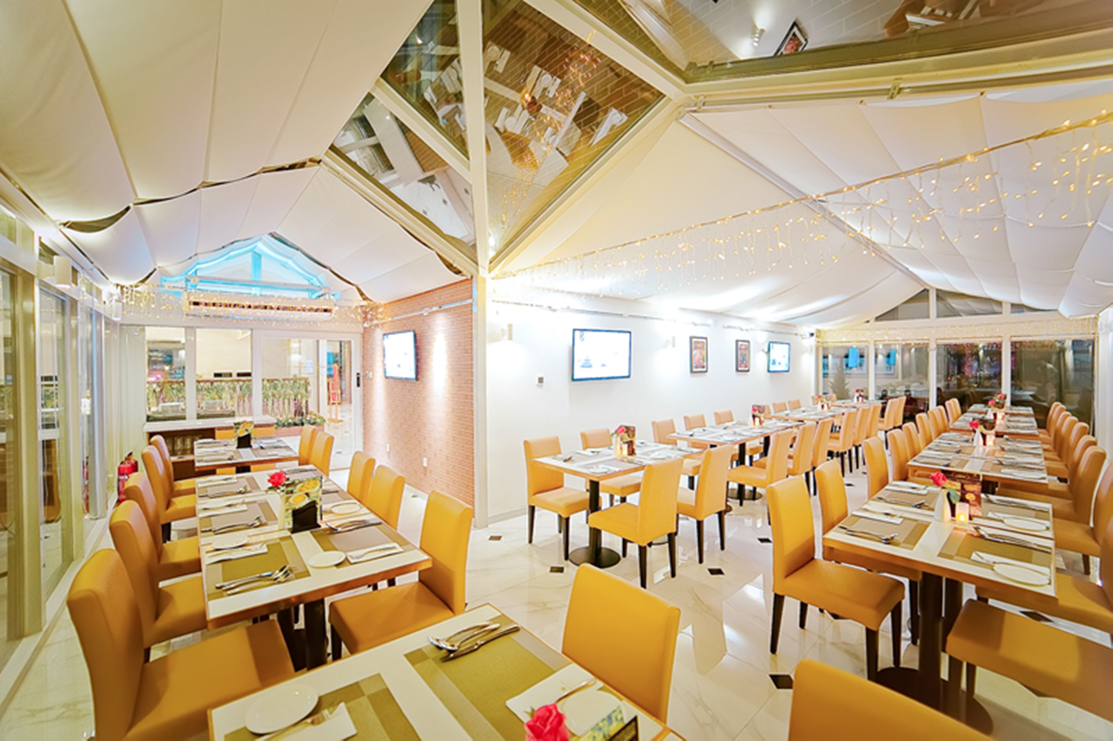 A restaurant with long tables and orange chairs, a high ceiling, and string lights.