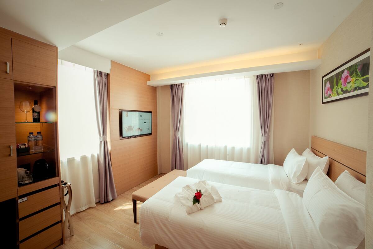 A hotel room with two beds, a wooden wardrobe, a television, and a window with curtains.