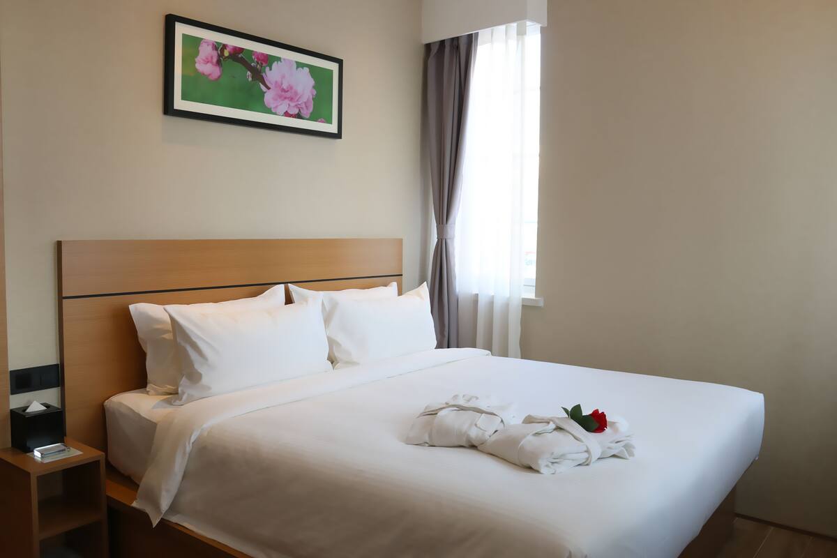 A neatly made bed with white linens, a wooden headboard, and a framed floral artwork above it.