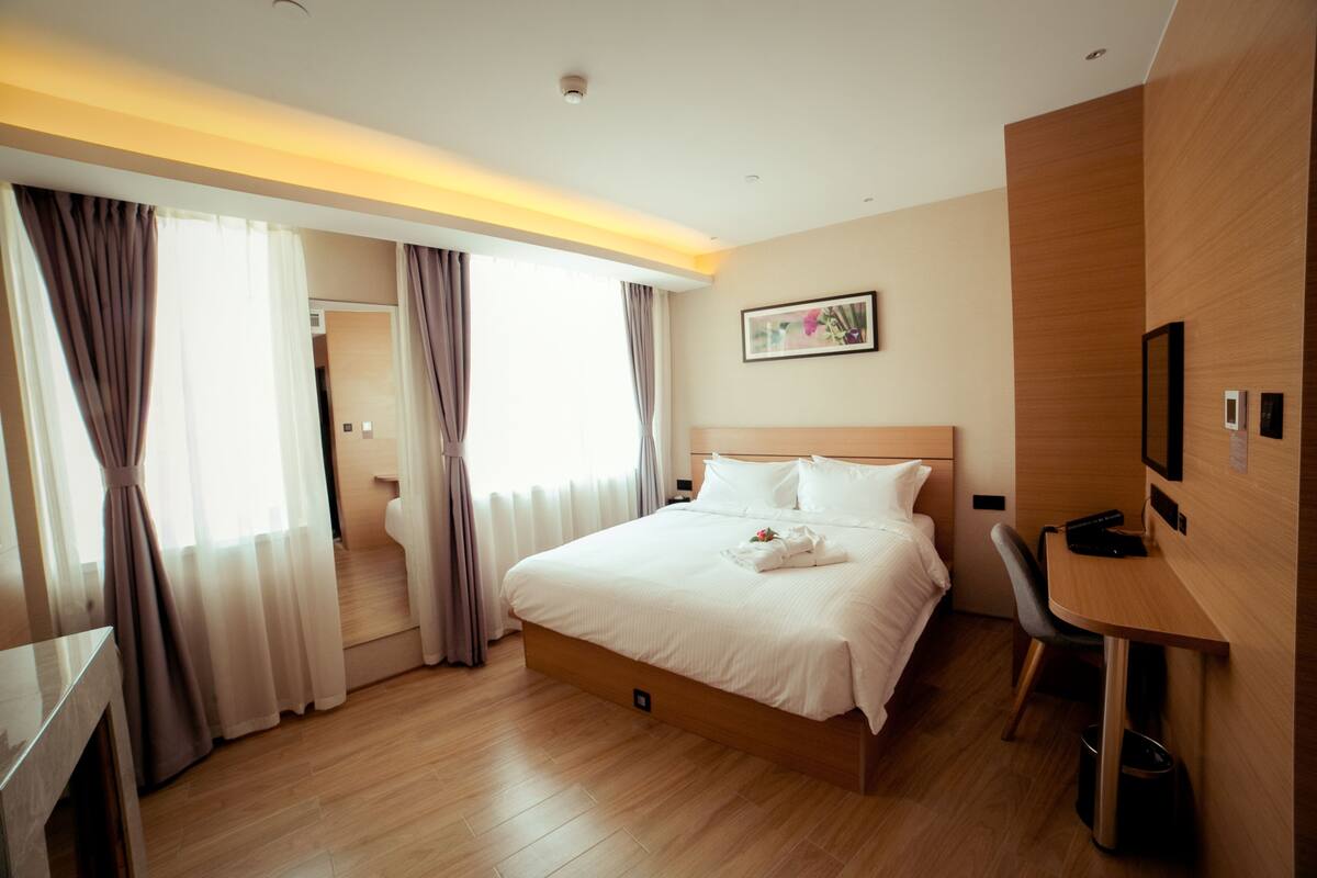 A hotel room with a bed, a desk, and a chair. There is a window with curtains, a picture on the wall, and a ceiling light.