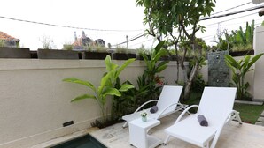 A poolside area with two white lounge chairs, a small table with a vase, and a stone wall with greenery. - Green Studio Apartment Sanur (Denpasar)
