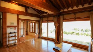 A spacious room with wooden flooring, a wooden table, a standing lamp, and large windows with curtains.