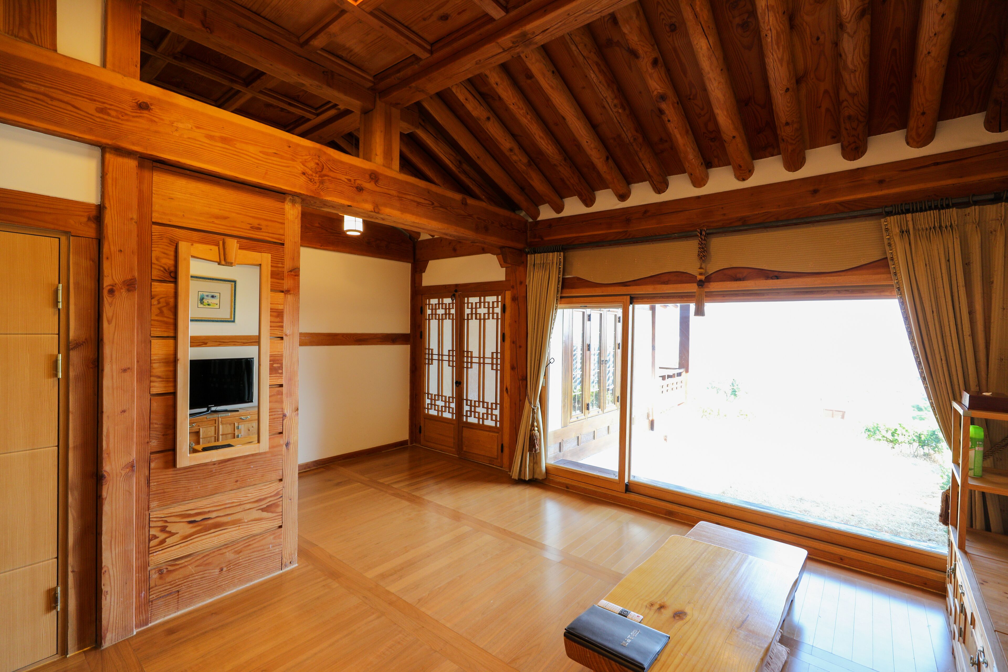A traditional Japanese room with wooden beams, tatami flooring, a television, sliding doors, and a view through large windows.