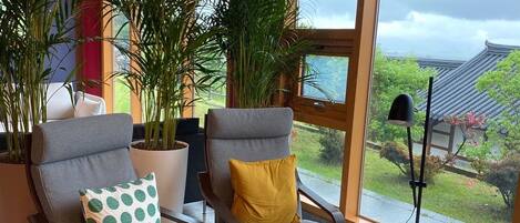 Un interior moderno con dos sillones grises, uno con un cojín de lunares verdes y el otro con un cojín amarillo, una mesita redonda y grandes ventanales que ofrecen vistas a arquitectura tradicional y vegetación.