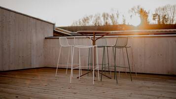 Una terraza de madera con una mesa y sillas, vista de árboles al fondo y un muro de madera.