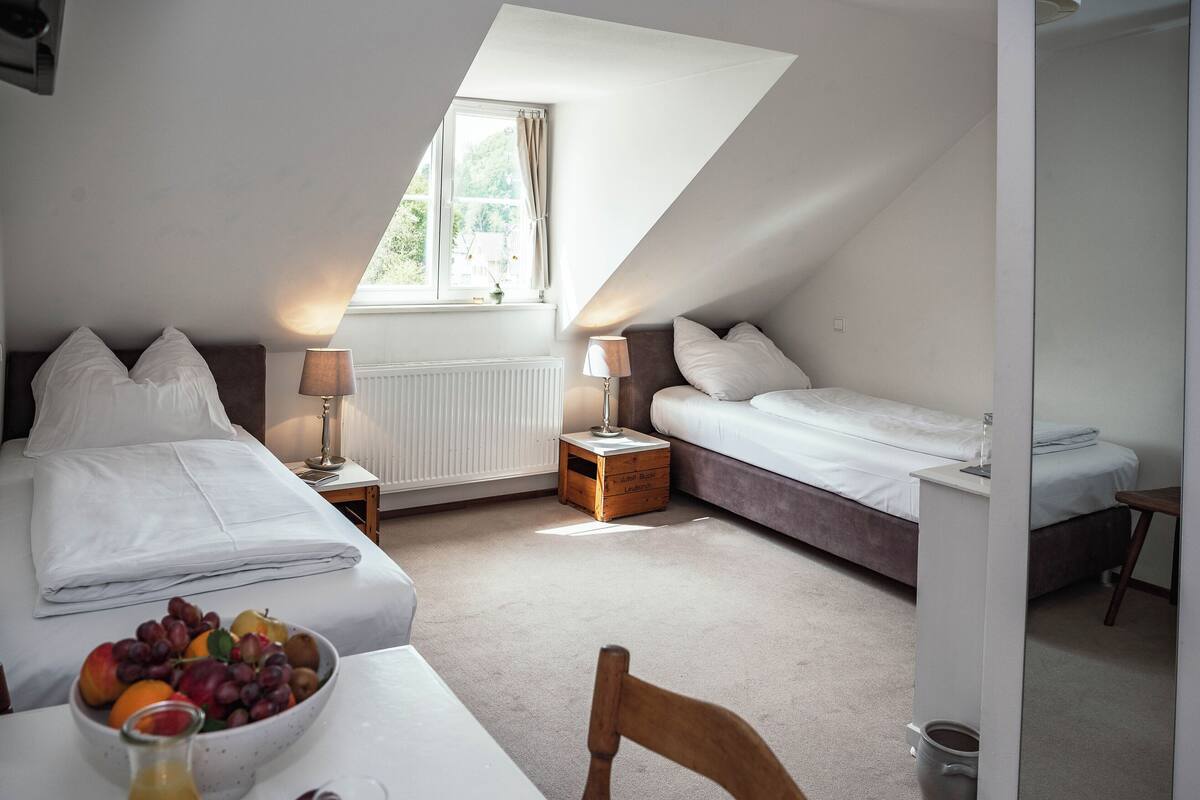 Une chambre avec deux lits, une fenêtre, une table de chevet avec une lampe et un bol de fruits sur une table.