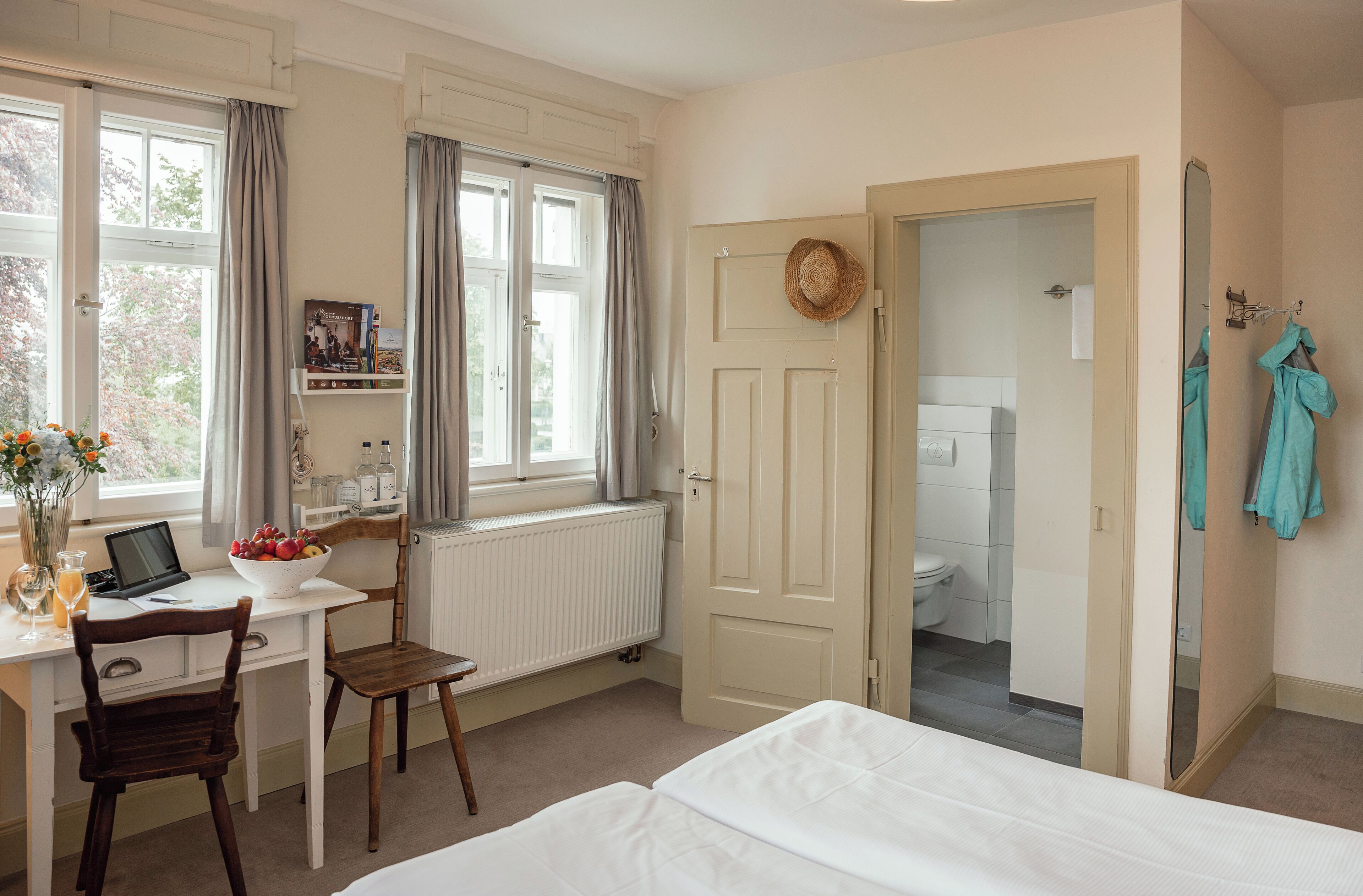 Un dormitorio con cama, escritorio con ordenador portátil, silla, ventana con cortinas y vista al baño.