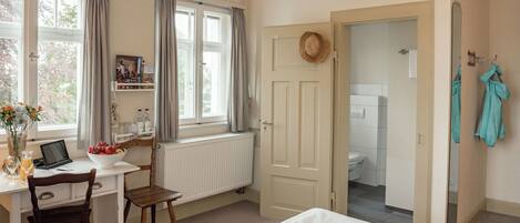 A bedroom with a bed, a desk with a laptop, a chair, a window with curtains, and a view of the bathroom.