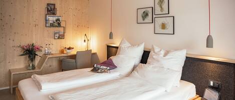 Una cama doble con ropa blanca, una mesita de noche con lámpara, un escritorio con silla, y cuadros en la pared.