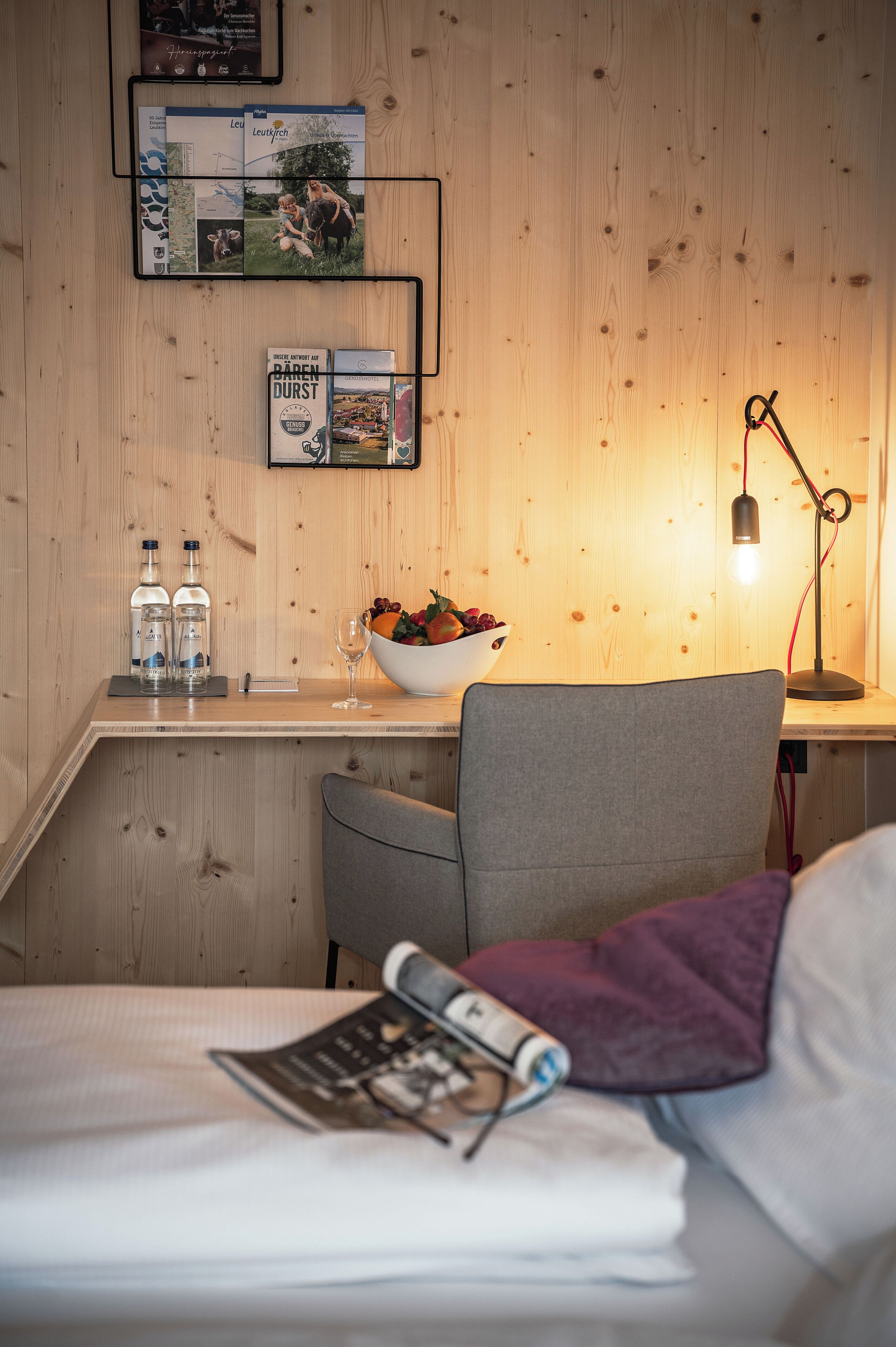 A hotel room with a wooden wall, a desk with a lamp, a bowl of fruit, and a bed with a magazine.