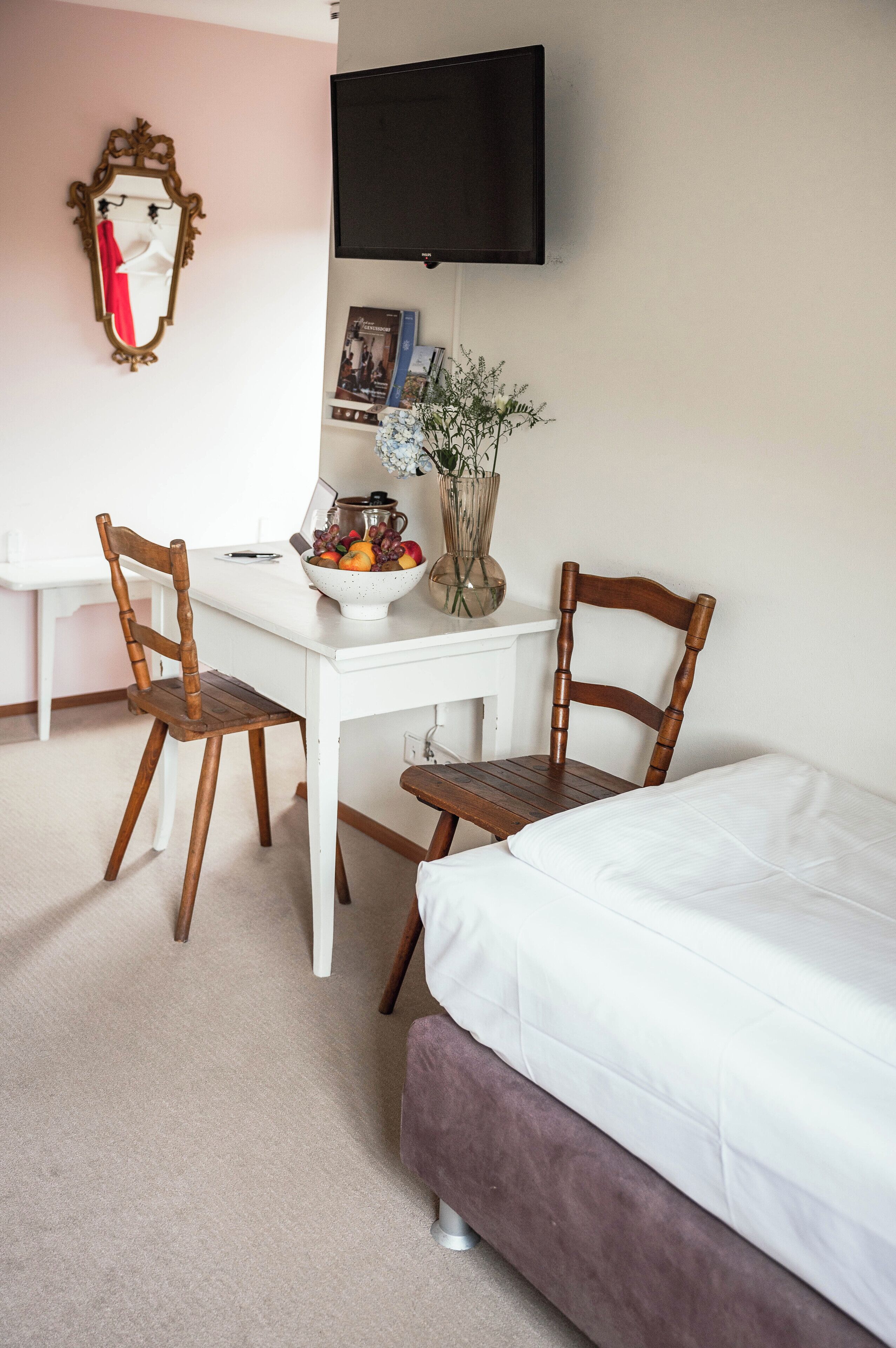 A hotel room with a white bed, a desk with a bowl of fruit, two wooden chairs, a television mounted on the wall, and a mirror with a red and white towel hanging on it.