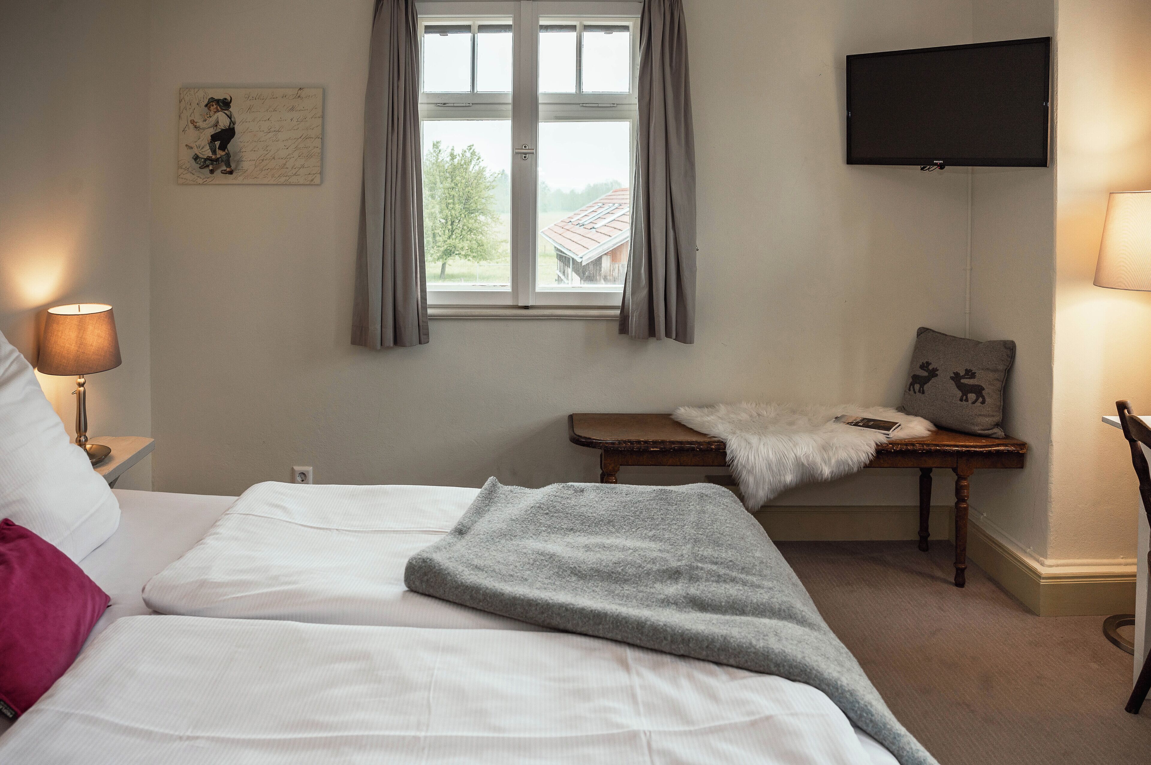 Un dormitorio con cama, una mesita de noche con lámpara, una ventana con cortinas, un televisor montado en la pared y un pequeño banco con una manta de piel.