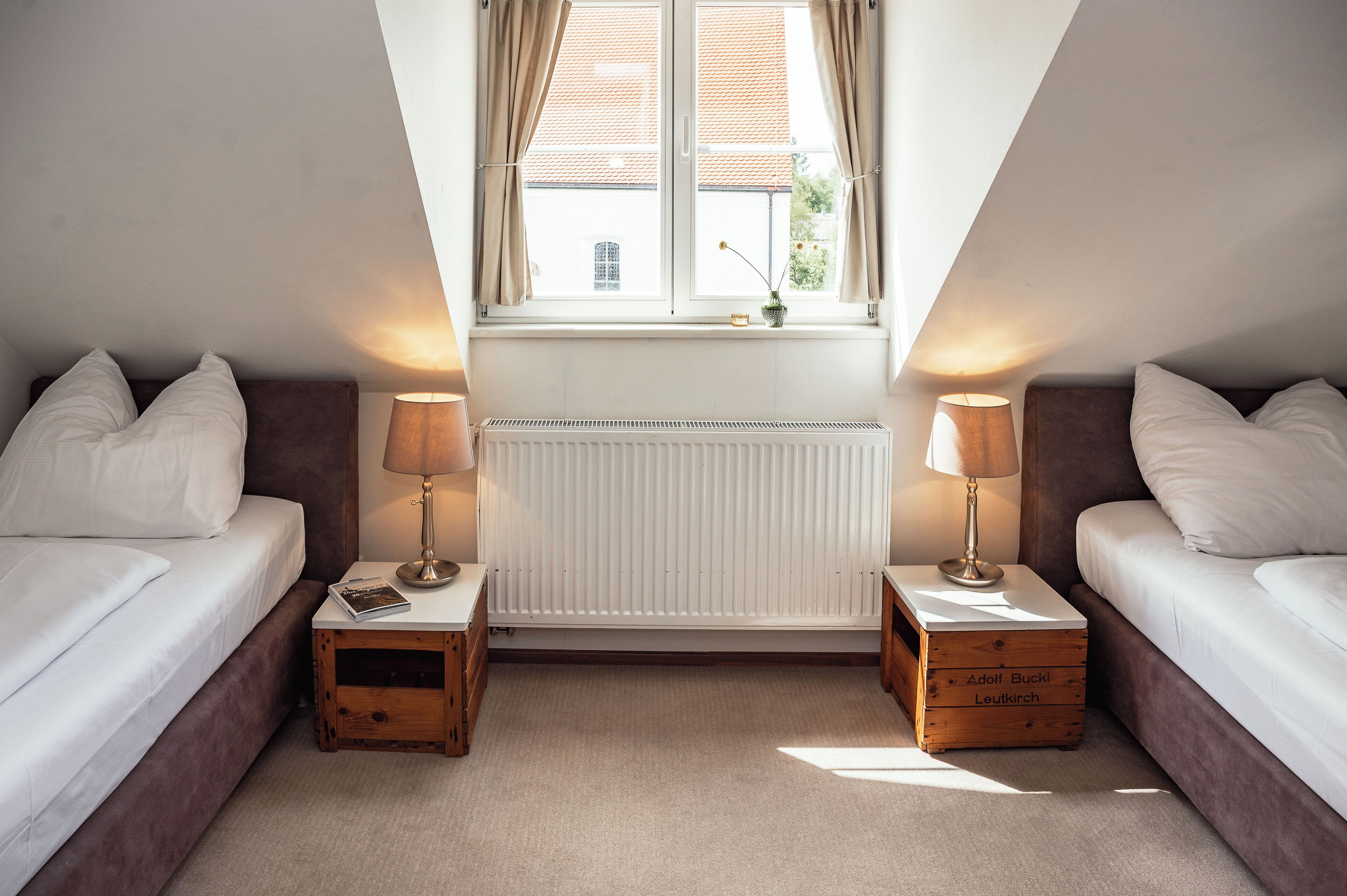 A bedroom with two beds, a window with curtains, a radiator, and bedside lamps.