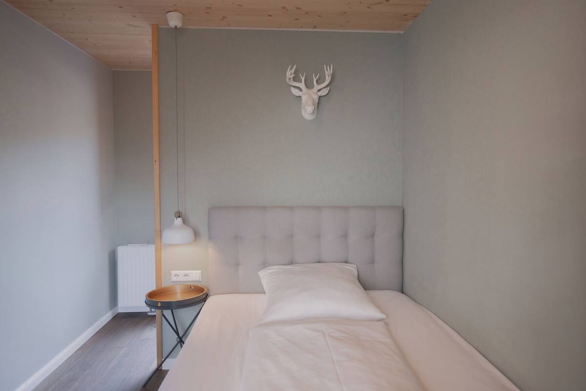 Une chambre à coucher comprenant un lit, une tête de lit, une décoration murale représentant une tête de cerf, une table de chevet avec une lampe et un radiateur.