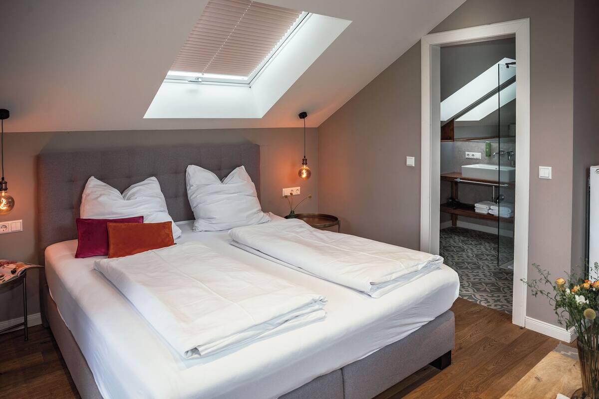 Une chambre avec une lucarne, un lit recouvert de draps blancs, une table de chevet avec une lampe, et une vue sur la salle de bain à travers une porte en verre.