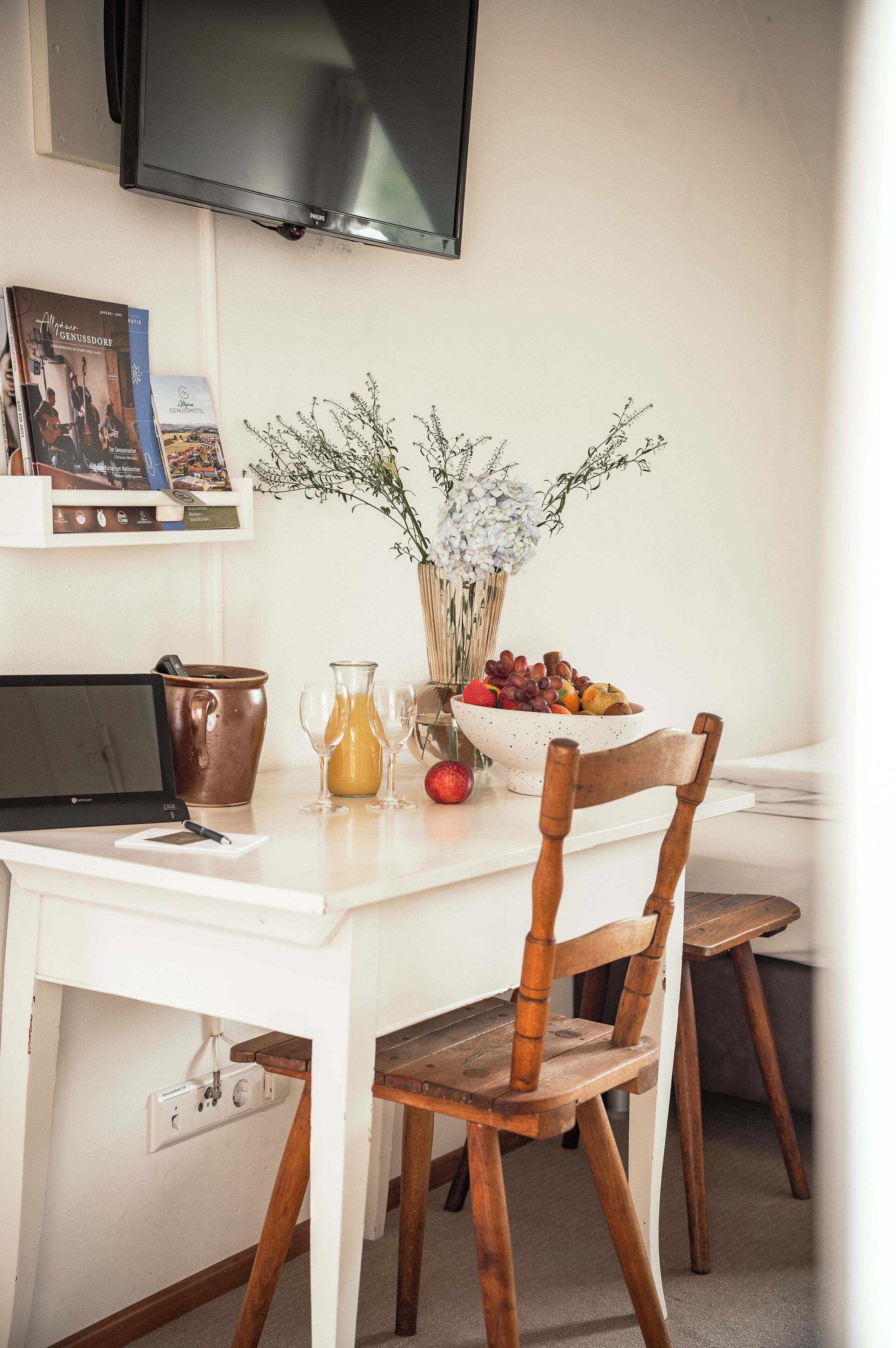 A dining area with a white table, a wooden chair, a laptop, a copper pitcher, a glass pitcher, a bowl of fruit, and a vase with flowers.