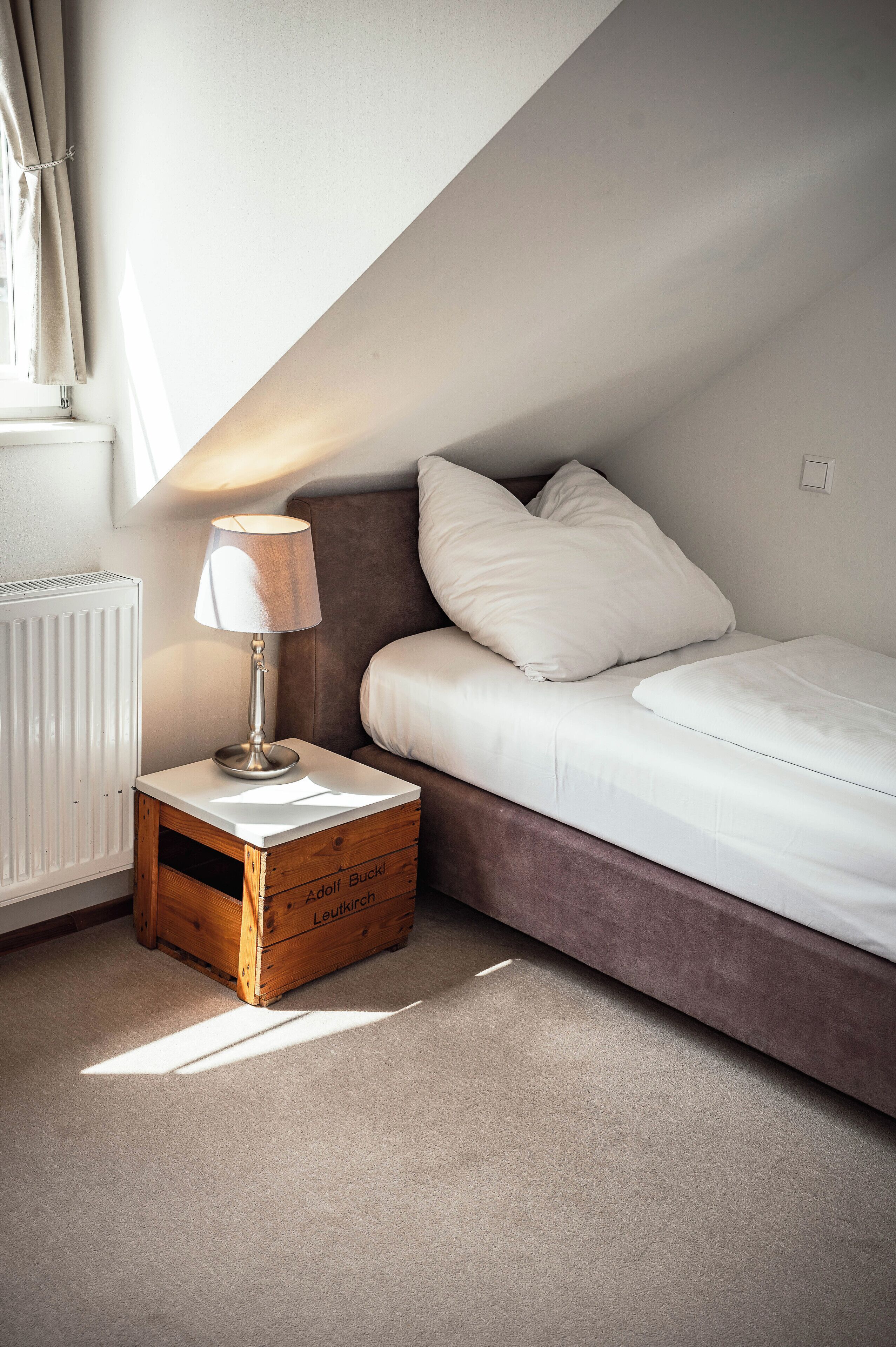 Un dormitorio con una cama individual, una mesita de noche con lámpara, un radiador y un baúl de madera.