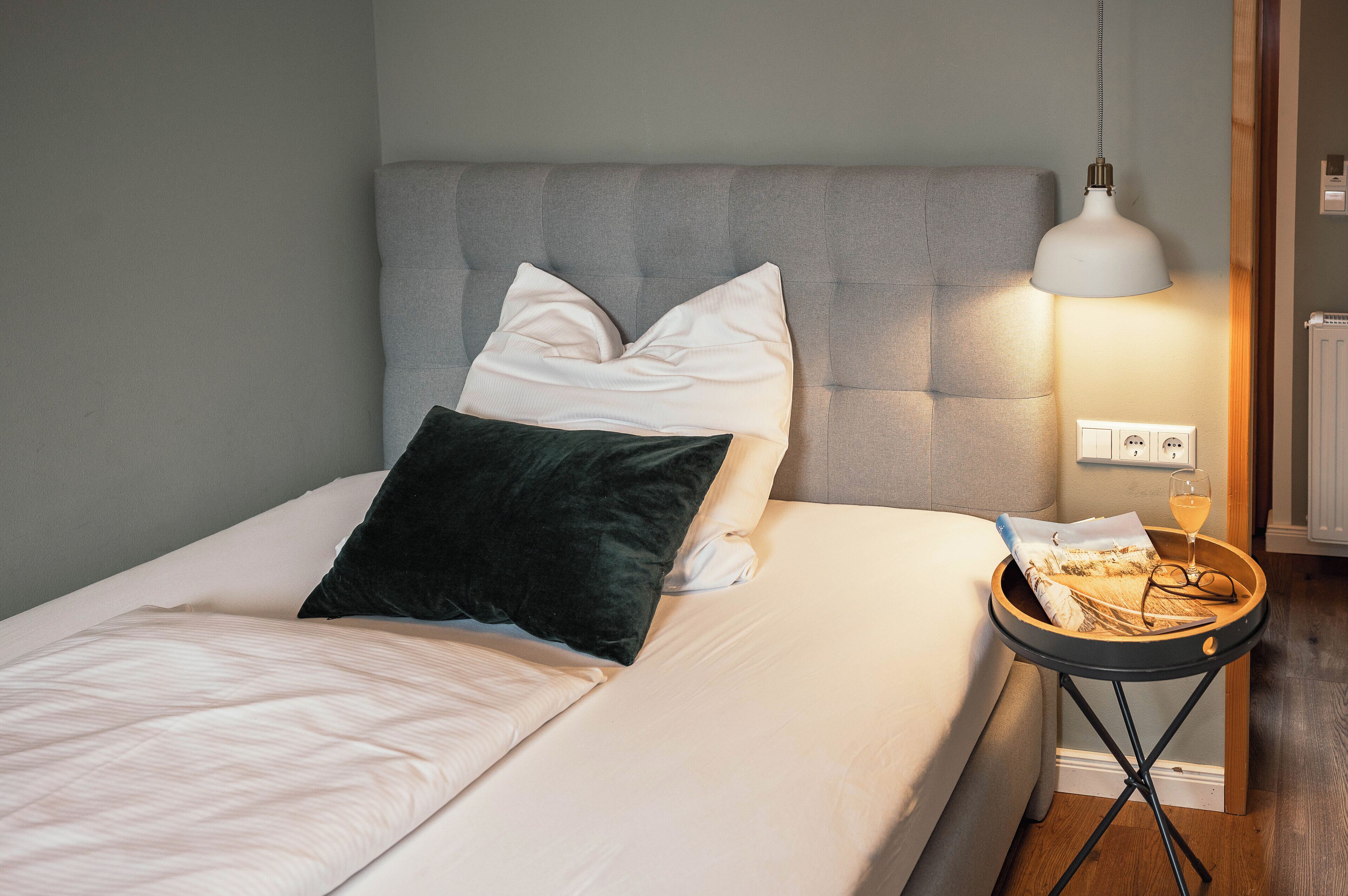 A neatly made bed with a grey upholstered headboard, a white pillow, a dark pillow, and a white comforter. There is a small wooden side table with a white lamp, a glass of wine, and a pair of glasses.