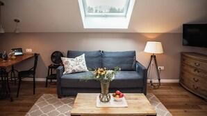 A living room with a dark blue sofa, a wooden coffee table with a vase of flowers and a fruit bowl, a lamp, and a TV mounted on the wall.