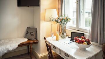 A cozy room with a white table, a laptop, a bowl of fruit, a lamp, and a window with curtains.