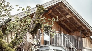A rustic wooden building with a staircase leading to a balcony, surrounded by trees and a fence.