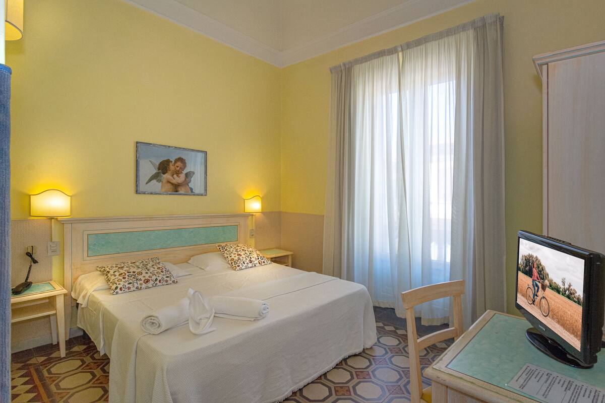 A hotel room with a bed, two bedside lamps, a television, and a window with curtains.