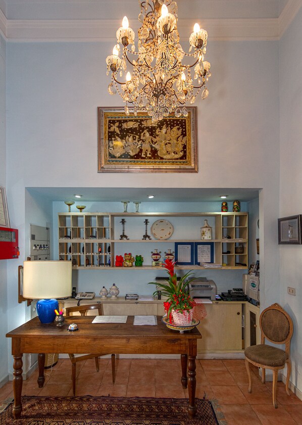 A dining area with a wooden table, chairs, a chandelier, a wall-mounted painting, shelves with various items, and a red fire alarm. - Hotel Arcangelo (Viareggio)