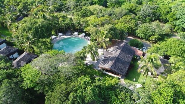Vista aérea de un resort con una piscina central rodeada de sillas de descanso, palmeras y edificios con techo de paja.