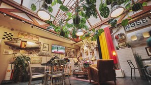 Interior de restaurante com tema vintage, móveis de madeira, teto alto com plantas penduradas e exposição de carros antigos.